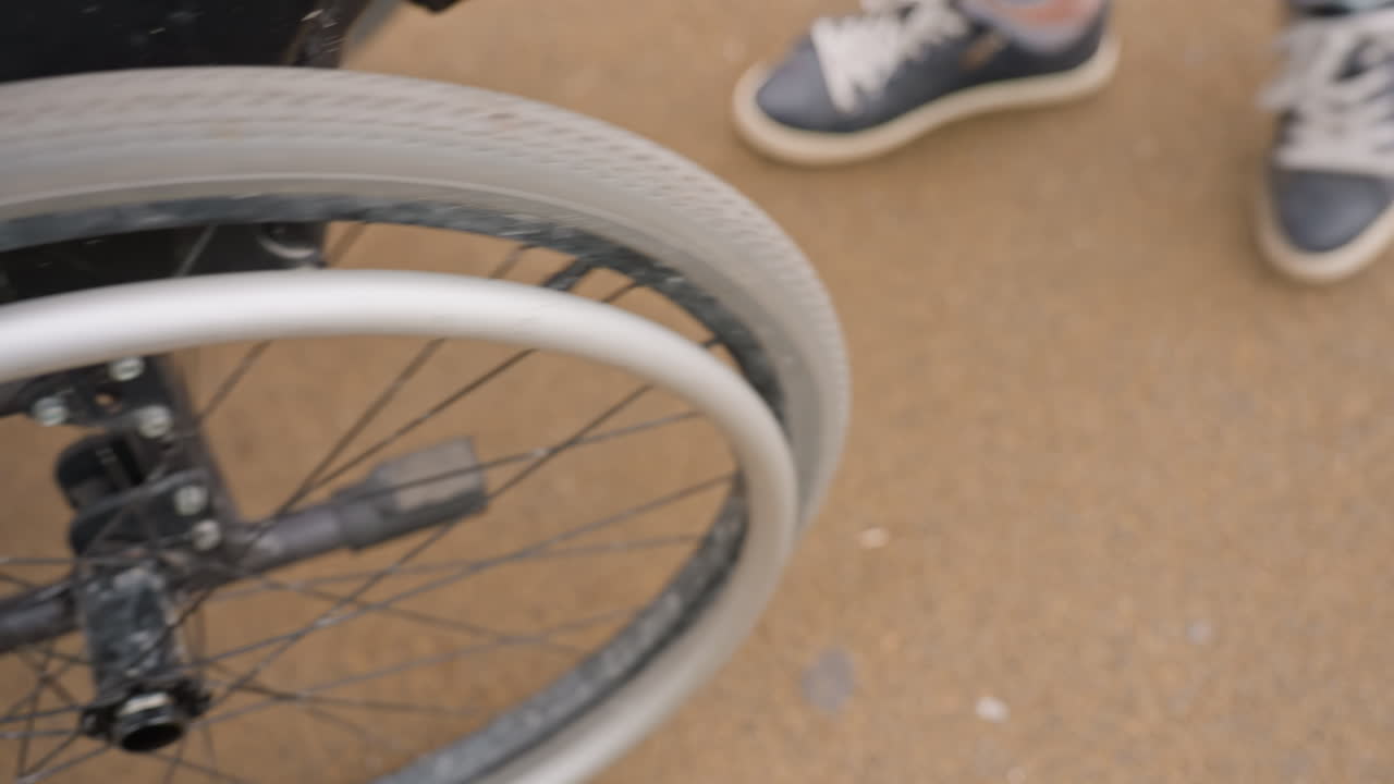 CloseUp Wheelchair Hand Adjusting Wheel Outdoors, White Woman In Black Jacket Grips Rim Near CompanionS Sneakers, Focus On Spokes, Caster And Brake, Textured Pavement, Practical Supportive