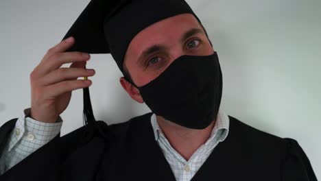retrato de feliz graduado poniéndose una máscara protectora, usando bata y gorra de graduación