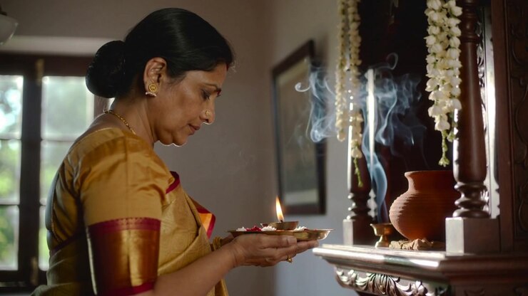 Holding brass thali with lit lamp, senior Indian woman in yellow saree offering prayer at shrine
