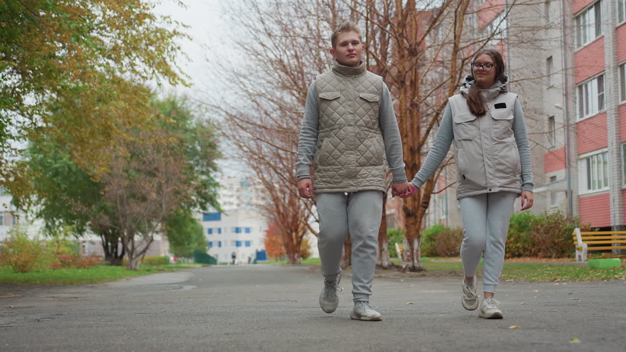 Evening stroll of young couple holding hands in matching outfits, walking along tree-lined sidewalk in peaceful urban area with blurred figure approaching in distance