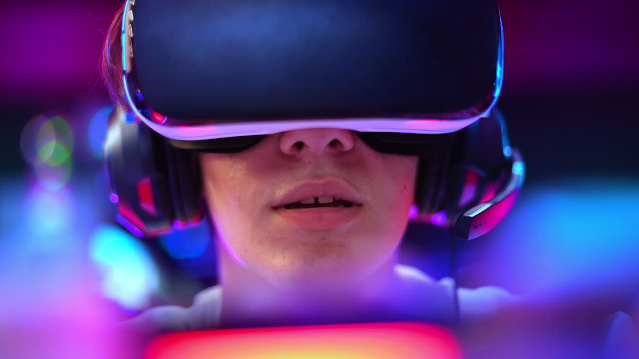 Primo piano di un adolescente che gioca a una console di gioco con visore VR e cuffie usando un gamepad. Illuminazione blu e rossa