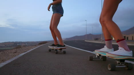 Dos-Amigos,-Vestidos-Con-Pantalones-Cortos,-Andan-En-Patineta-Por-Una-Carretera-Durante-El-Atardecer,-Con-El-Telón-De-Fondo-De-Montañas-Y-Un-Hermoso-Cielo,-Todo-Capturado-En-Cámara-Lenta.