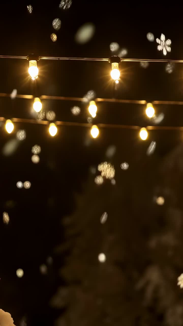 Vertical video: Snowing harder, warm string lights glowing, flakes drifting in yard night near tree