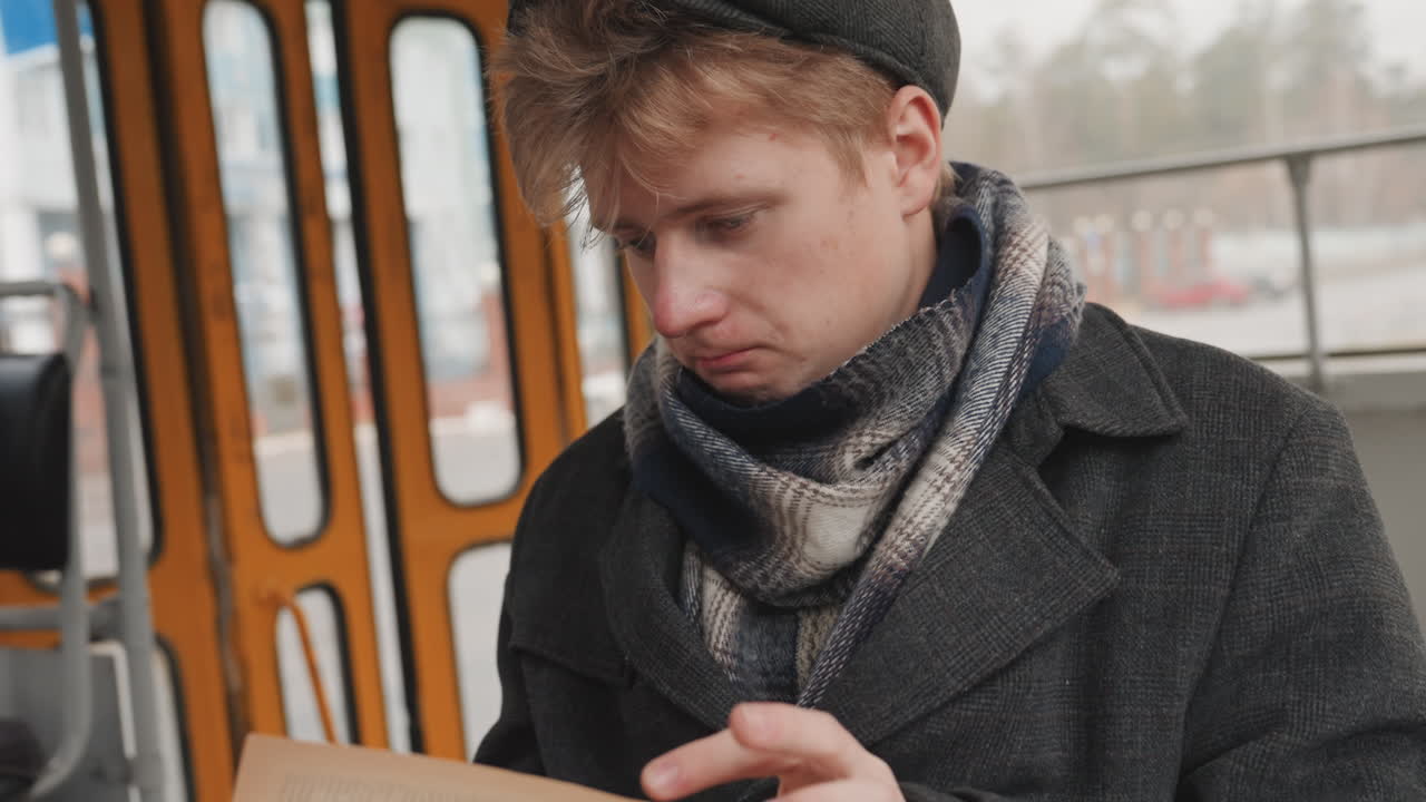 Weißer Mann prüft Notizen auf einem Straßenbahnsitz, trägt Schal und Baskenmütze. Nachdenklicher junger Mann notiert Ideen in sein Notizbuch neben einem regennassen Fenster. Vintage-Interieur und vorbeiziehende Uferpromenade prägen die gedämpfte, bewölkte Stimmung.