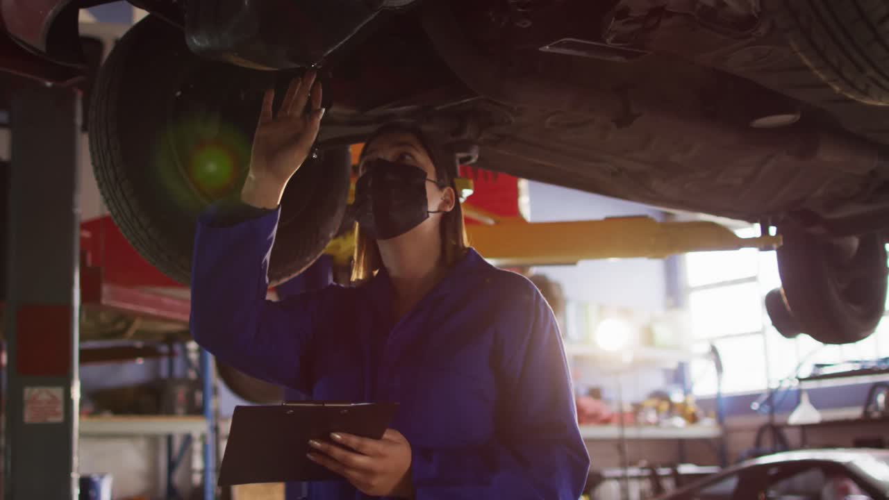 mecánico femenino con máscara facial tomando notas en el clipboard debajo de un coche en una estación de servicio de coches