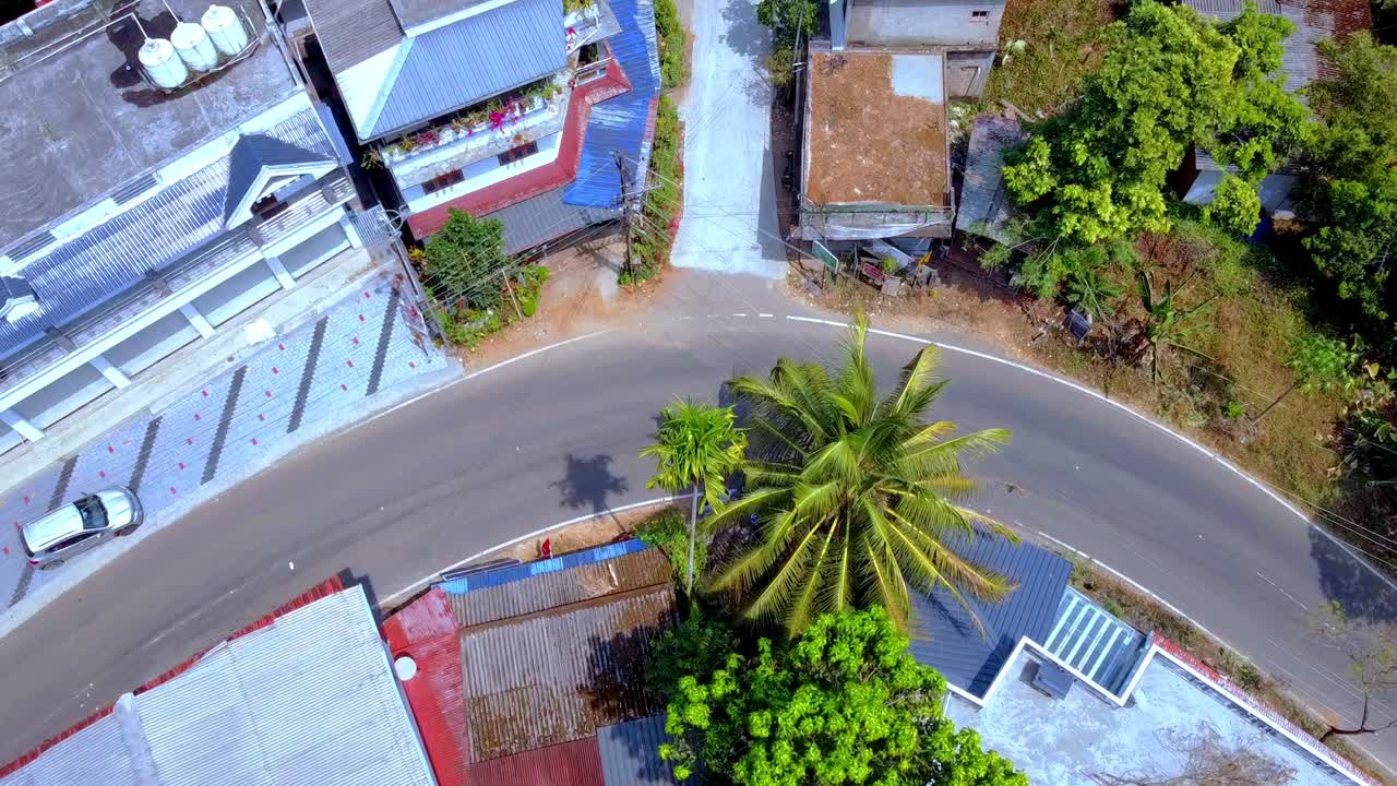 Ripresa aerea con drone dall'alto di una strada asfaltata tortuosa in un lussureggiante villaggio tropicale. Scooter e auto percorrono una curva stretta circondata da case colorate e palme