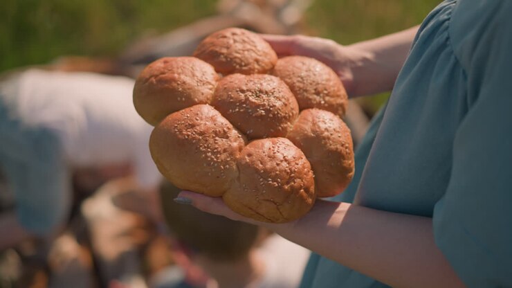 A woman wearing a blue dress holds a cluster of fresh sesame seed bread rolls, with her children blurred in the background. The close-up shot focuses on the bread