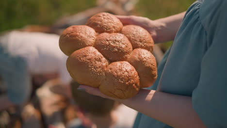 a woman wearing a blue dress holds a cluster of fresh sesame seed bread rolls, with her children blurred in the background. the close-up shot focuses on the bread