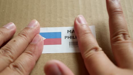 Hände-Bringen-Das-Flaggenetikett-„Made-In-Philippines“-Auf-Einem-Versandkarton-Mit-Einem-Barcode-In-Premiumqualität-An