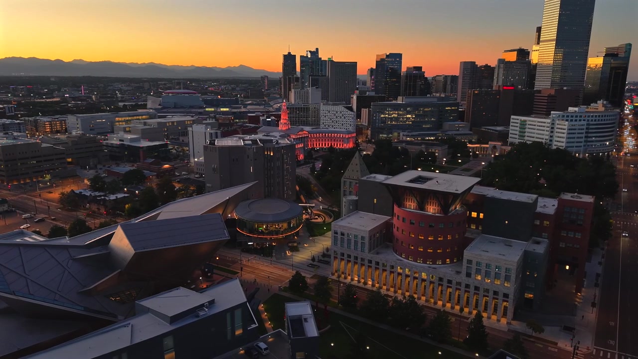 Un drone scende e orbita il Denver Art Museum al tramonto, catturando colori intensi nel cielo insieme alle luci notturne luminose del centro di Denver sottostante