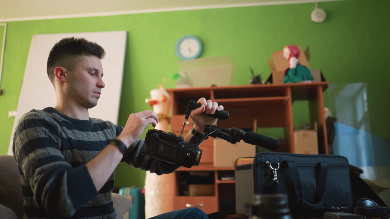 Mann justerer kamera med stativ og mikrofon i innendørs studio, fokusert på innstillinger og utstyr. Grønne vegger, klokke og hylle med dekorative gjenstander i bakgrunnen. Forbereder videoproduksjon