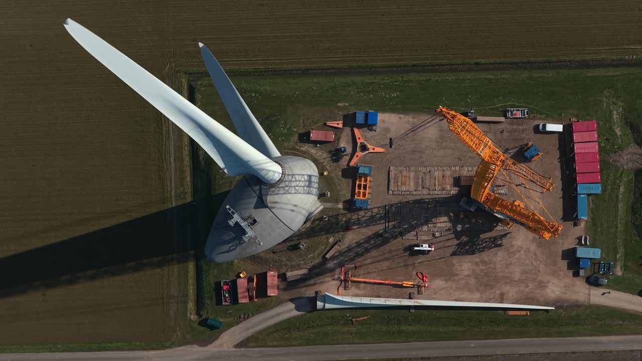 Vista aérea cenital de una obra mostrando una grúa que eleva y monta una pala gigante en el buje de un aerogenerador para la producción de energía verde.
