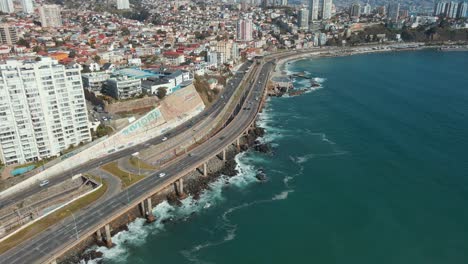 Muñeca-Aérea-En-El-Tráfico-En-La-Avenida-Junto-A-La-Costa-Del-Mar-Turquesa,-Edificios-De-La-Ciudad-De-Viña-Del-Mar-Y-Colinas-En-La-Parte-Trasera,-Chile