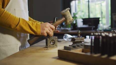 midsection of caucasian female jeweller in workshop wearing apron, using hammer, making jewelry