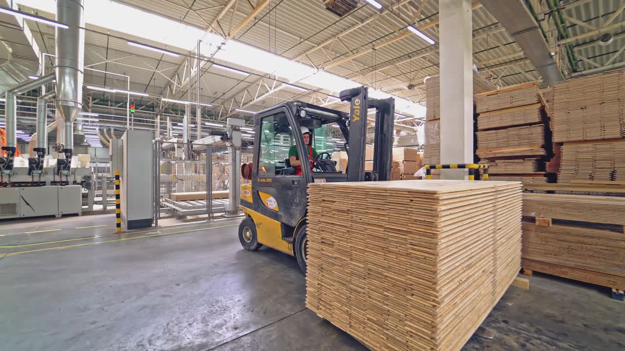 An operator in a white helmet works on an electric loader and carries it a pile of wooden parquet boards to another place in the warehouse. New plant for the manufacture of parquet.