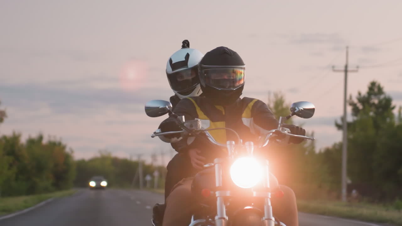 Close up of motorcycle rider with passenger cruising along road at sunset, headlight glowing bright, distant car headlights visible against greenery