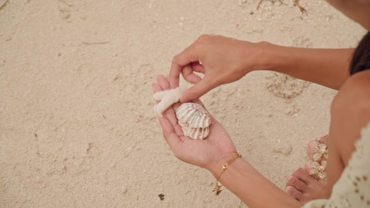 Beach closeup womans hands arranging shells into small keepsake on sunlit sand, detailed craft motion, delicate placement of scallop and coral, bracelet on wrist, intimate creative ritual, seaside