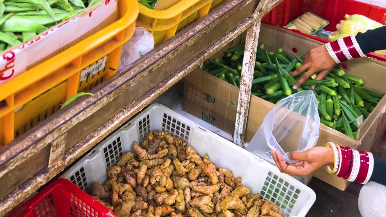 A person wearing striped sleeves picks fresh turmeric roots and places them into a plastic bag at a vibrant outdoor vegetable market under bright natural lighting