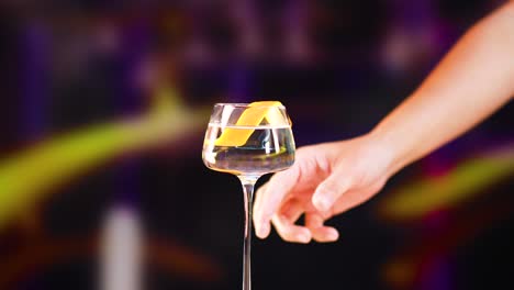 a hand reaches for a martini glass with a lemon twist against a colorful, blurred background