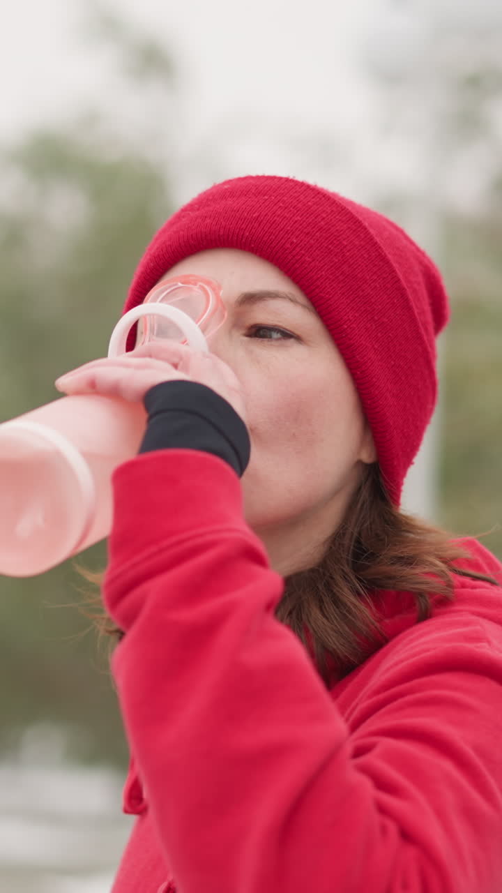 은 베니와 후디를 입은 여자가 은 겨울 환경에서 초록색과 철 철로로 흐릿한 배경에 분홍색 병을 사용하여 야외에서 물을 마시고 있습니다.