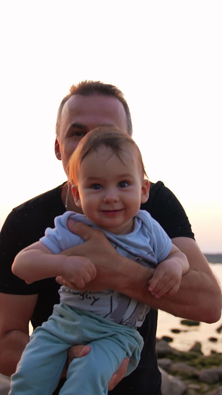 Loving father with cute little toddler in his arms approaching camera. Adorable baby boy is laughing cheerfully. River at backdrop. Vertical video