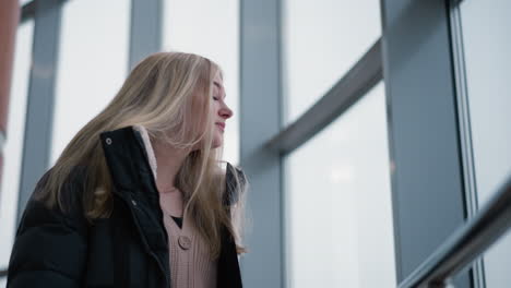 young lady gazing thoughtfully through glass building window, her long hair cascading down her jacket as she looks back contemplatively, immersed in a moment of reflection in an urban setting