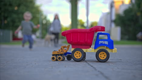 unrecognizable young boy running from his mother towards the camera to play with some toy trucks placed on the street