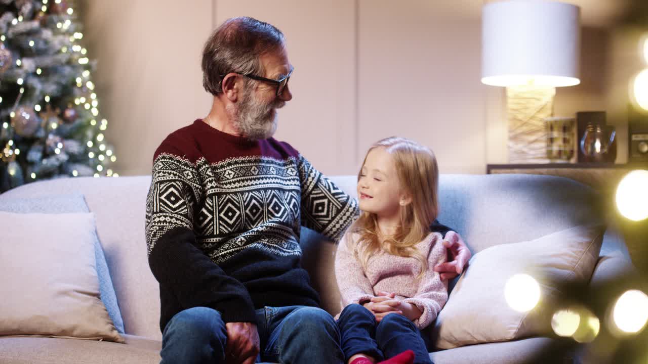 feliz abuelo cariñoso con anteojos pasando tiempo junto con una linda nieta sentada en una casa decorada cerca del brillante árbol de navidad y abrazándose