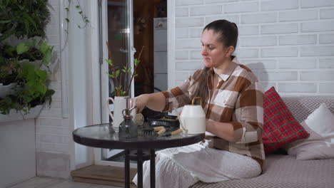 nature enthusiast seated on sofa beside indoor garden sprinkles seeds into plastic plate on glass table surrounded by watering can gardening tools cuttings cushions nurturing ritual indoors