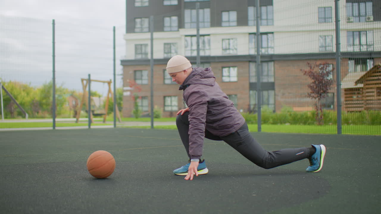 white senior man stretching on court, urban apartment complex backdrop, wearing jacket and knit cap, performing lunges and ground reach beside basketball on wet asphalt, cool morning atmosphere