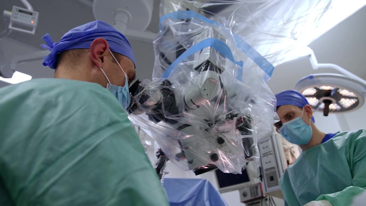 Focused male surgeon looking at microscope binoculars. Assistant stands on the opposite. Low angle view.