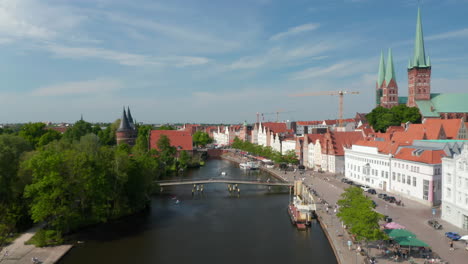 Forwards-fly-above-Trave-river-flowing-around-medieval-historic-city-centre.-Wide-waterfront-restaurant-terraces-and-people-walking-along-river-Luebeck,-Schleswig-Holstein,-Germany