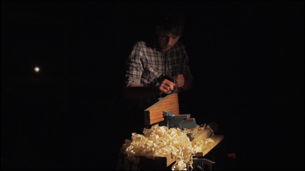 빛 속으로 걸어가는 목수의 설정 샷은 소나무 목재를 손으로 대패질하기 시작합니다.