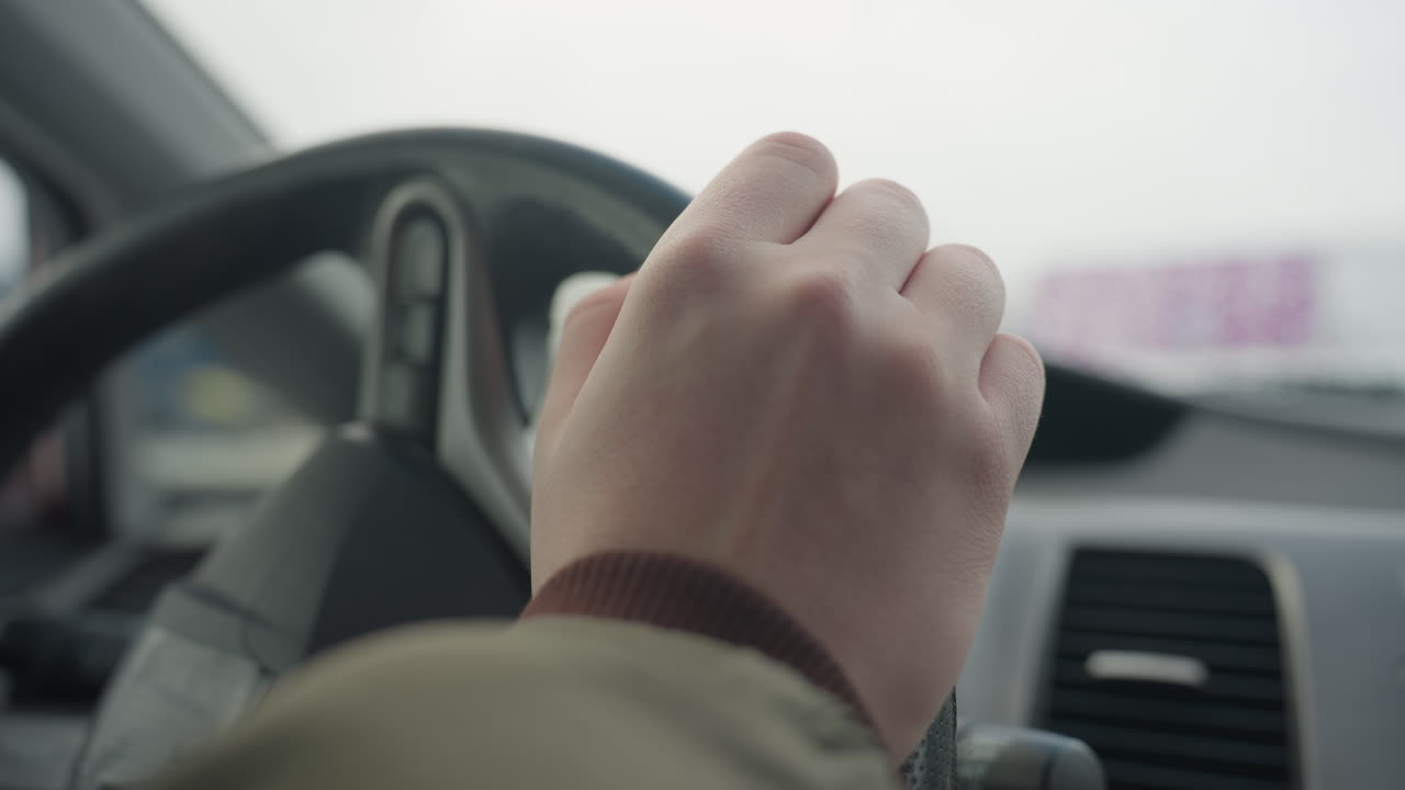 close up of fair hand gently tapping steering wheel, person wearing winter jacket, natural daylight coming through window with blurred snowy background and dashboard details visible