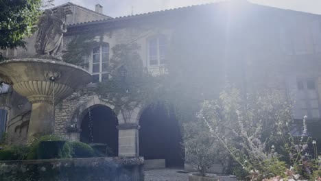 Wunderschönes-Altes-Haus-Mit-Verspielten-Bögen,-Bedeckt-Mit-Hängenden-Pflanzen,-Mit-Einem-Kleinen-Romantischen-Brunnen-Im-Vordergrund-Bei-Fantastischem-Wetter-In-Einem-Kleinen-Hirtorich-Dorf-In-Frankreich