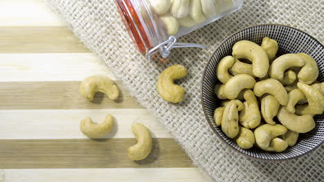 Panning-shot-across-striped-wooden-table,-Hessian-cloth-glass-jar-ceramic-bowl-filled-with-halved-cashew-nuts