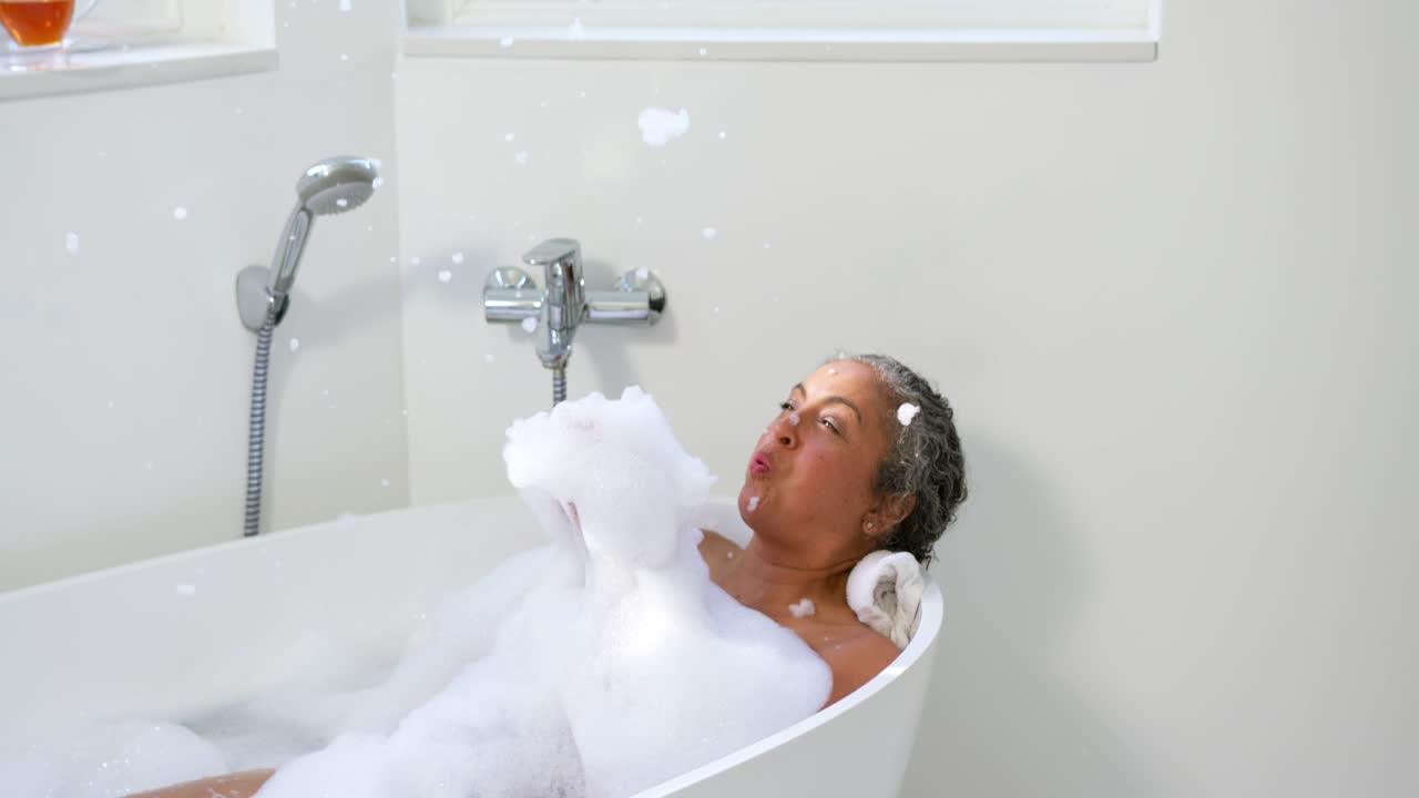 Senior female reclining in bathtub on rolled towel, scooping foam and blowing bubbles, relaxing