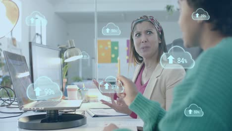 Cloud-icons-uploading-data-over-diverse-female-colleagues-discussing-at-computer-in-office