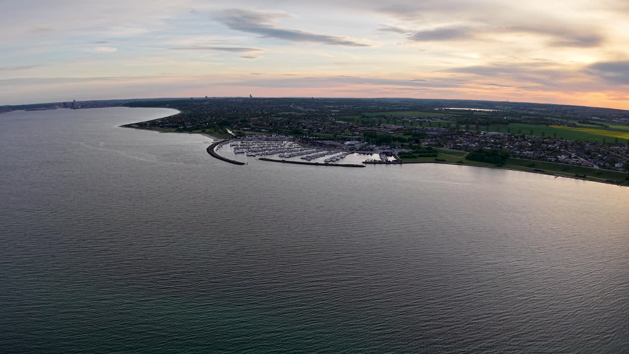 A drone flies over a calm body of water during sunset, showing a coastline with greenery and a harbor in Aarhus Bay, Denmark