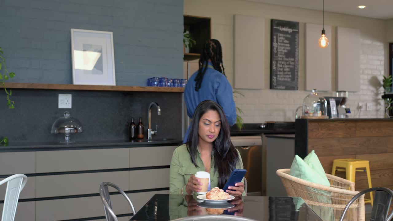 Mujer india de mediana edad sentada en una cafetería revisando el teléfono antes de que el barista coloque café y un pastel