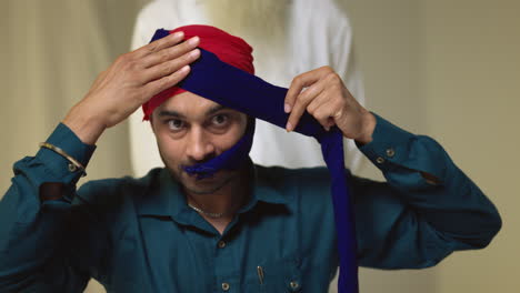 Fotografía-De-Estudio-De-Un-Hombre-Sikh-Mayor-Que-Ayuda-A-Un-Hombre-Sikh-Más-Joven-A-Atar-La-Tela-Para-Un-Turbante-Contra-Un-Fondo-Liso,-Filmada-En-Tiempo-Real-4