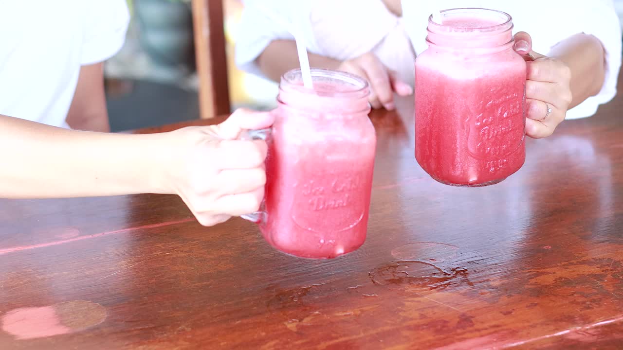 Hands clink watermelon juice glasses at a wooden table in a sunny Phuket cafe, capturing a moment of joy and refreshment