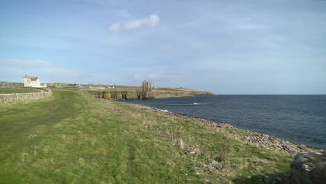 Aerial-shot-moving-across-the-coastline-towards-Keiss-Castle-with-an-old-WW2-bunker-in-the-foreground-on-a-sunny-day,-Caithness,-Scotland