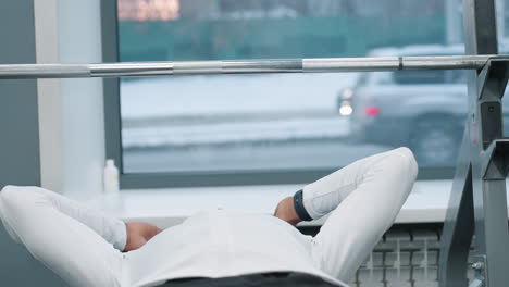 close up of athlete adjusting headphones while lying on bench press under chrome barbell window behind with car passing outside rubber floor gym equipment visible focused pre workout setup