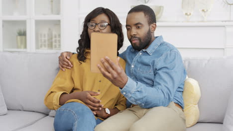 Young-Couple-Sitting-On-Couch-In-Living-Room-And-Videochatting-On-Tablet-Device