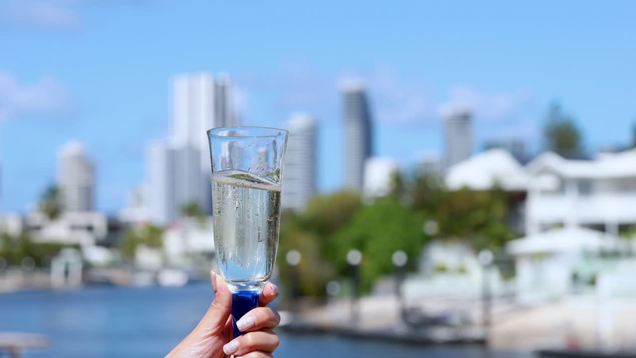A hand swirls a champagne glass, causing liquid motion, against a sunlit urban waterfront with modern buildings and blue sky in the background