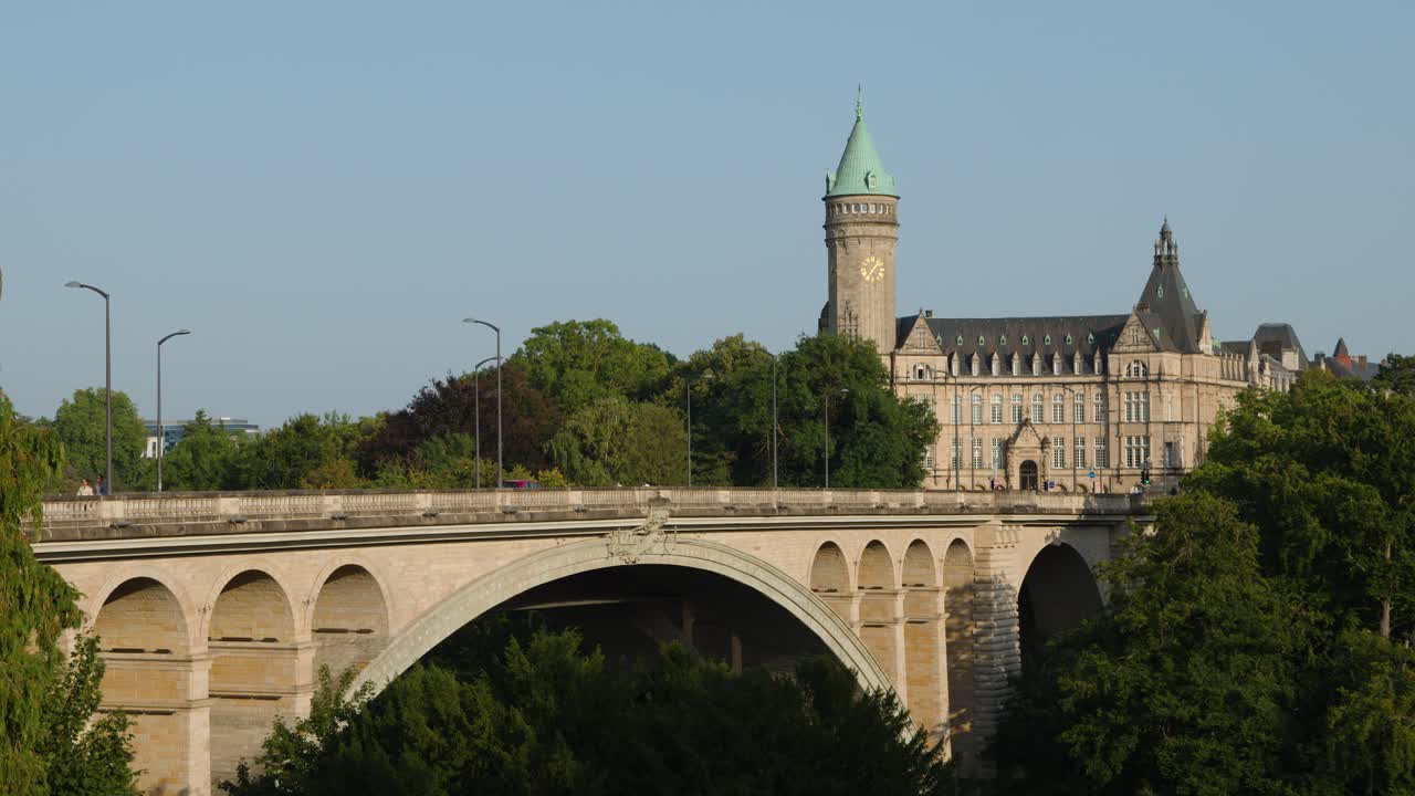 Smooth lateral pan reveals arched stone bridge, clock tower, and green-roofed building under clear sky