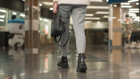 low angle view of mall goer stepping off moving walkway onto glossy tiled hall floor wearing light denim pants and chunky black boots under bright overhead lights in modern retail corridor