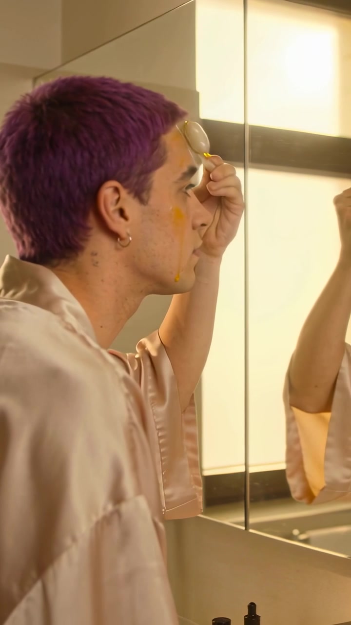 Video verticale: Uomo in accappatoio si applica un siero con un rullo su fronte e guancia davanti allo specchio del bagno.