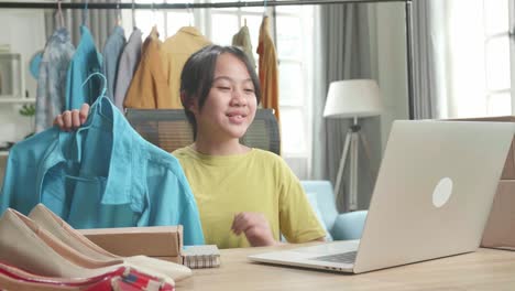 asian young girl online seller recommending a shirt while using computer for selling clothes at home